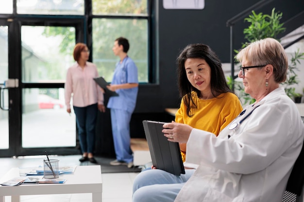 Senior doctor and patient attending consultation appointment in hospital waiting area, filling medical report using tablet computer, discussing about healthcare. People at checkup visit.