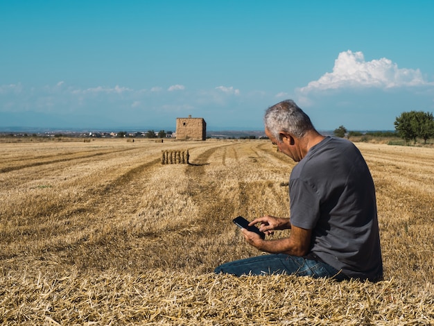 Senior farmer sitting on straw bale looking at mobile phone in field Agriculture concept