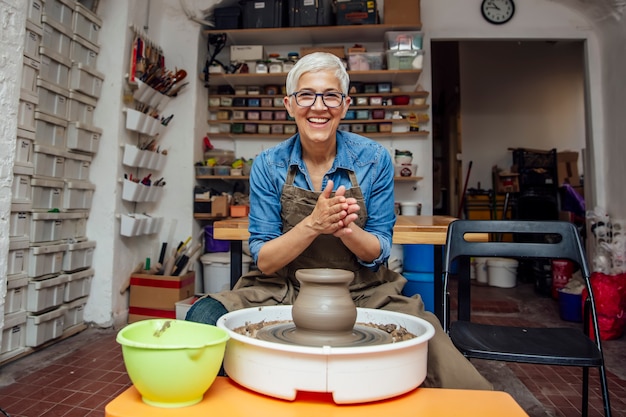 Senior female potter working on pottery wheel while sitting  in her workshop