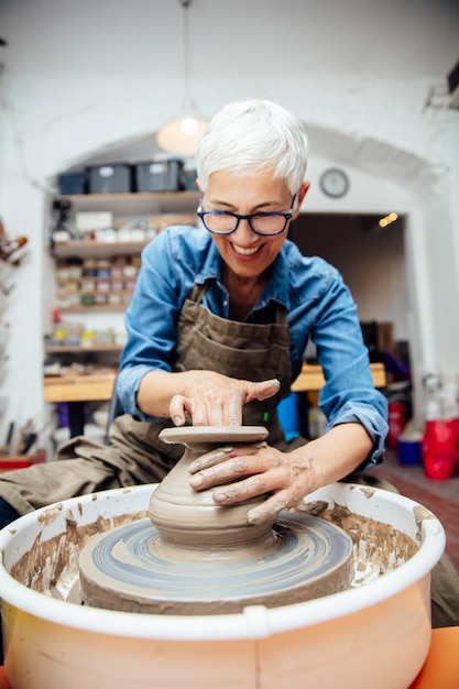 Senior female potter working on pottery wheel while sitting  in her workshop