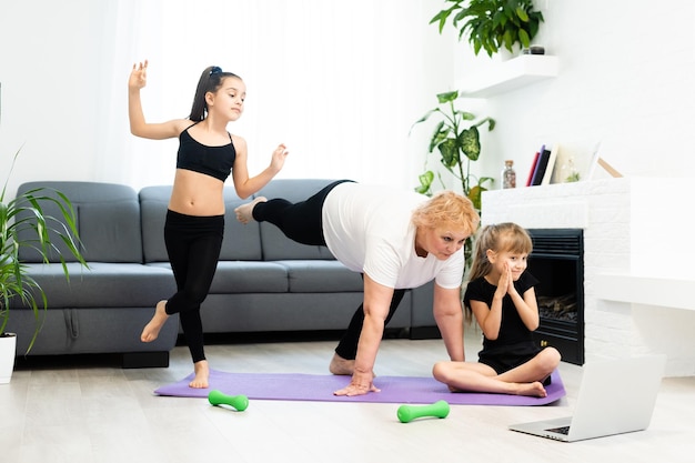 senior grandmother workout yoga withgranddaughters preschooler. They are sitting on mat at cozy home interior. Sport, parenthood and people concept.