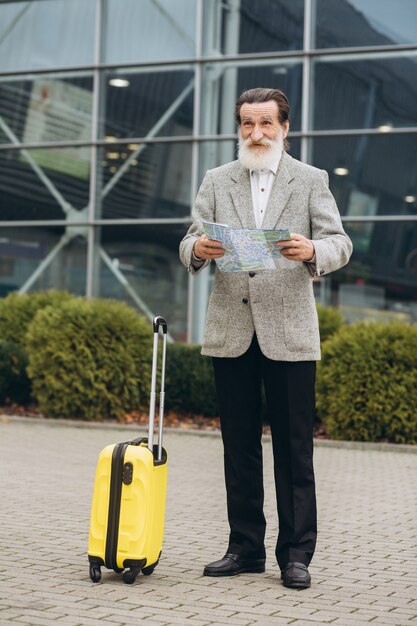 Senior gray-bearded man with carrying suitcase and city map is walking along the airport building. He is looking aside pensively. Copy space on right side