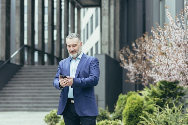 Senior grayhaired man a businessman in a suit stands near the modern office center and calls a taxi