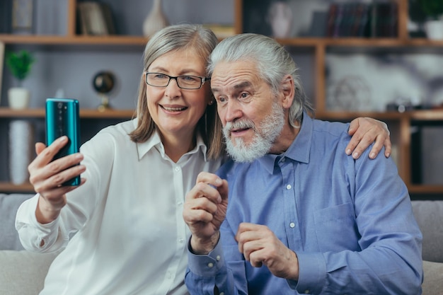 Senior grayhaired married couple man and woman at home together play video call sit on sofa at home use phone for call and online meeting