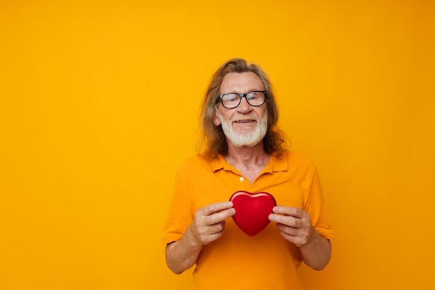 Senior greyhaired man yellow tshirt and glasses posing heartshaped box isolated background