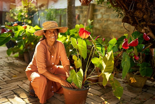 Photo senior happy indian female farmer in straw hat sitting in garden taking care of potted plants elderly sri lankan smiling woman in her yard planting flowers in pots active senior gardening concept