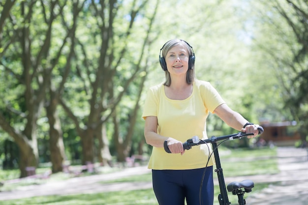 Senior happy woman on a walk in the park with a bicycle listens to music on headphones and walks on a summer day