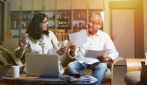 Senior Indian asian couple accounting, doing home finance and checking bills with laptop, calculator and money also with piggy bank while sitting on sofa couch or table at home