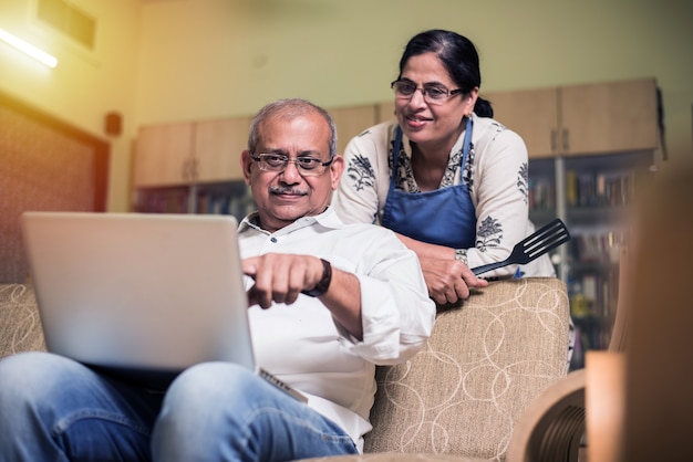 Senior Indian asian couple accounting, doing home finance and checking bills with laptop, calculator and money also with piggy bank while sitting on sofa couch or table at home