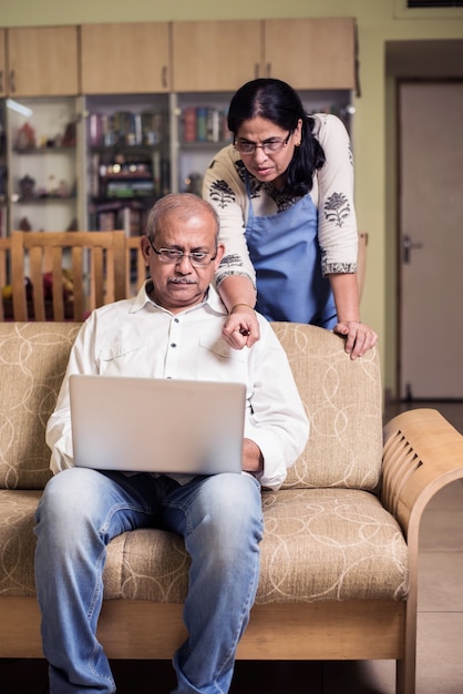 Senior Indian asian couple accounting, doing home finance and checking bills with laptop, calculator and money also with piggy bank while sitting on sofa couch or table at home