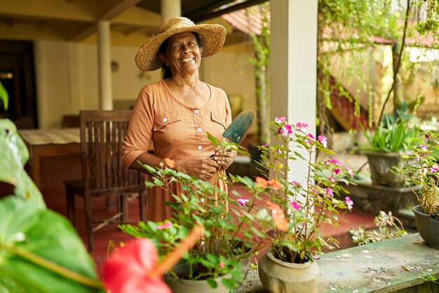 Photo senior indian female farmer in straw hat with gardening scoop in garden elderly sri lankan happy woman standing among flowers and plants smiling in orchard farming and gardening concept