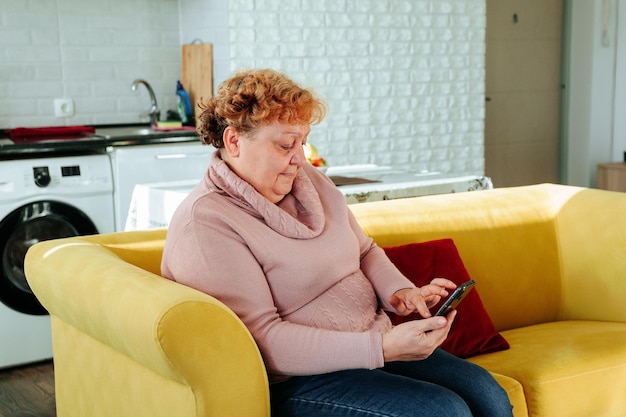 Senior lady using smartphone surfing the net sit on couch at home in the living room older fat peopl...