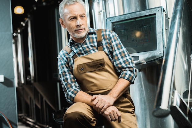 Senior male brewer in working overalls sitting in brewery