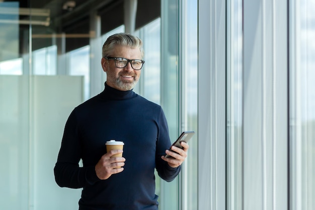 A senior male businessman stands in an office center near a window and uses the phone he holds a cup