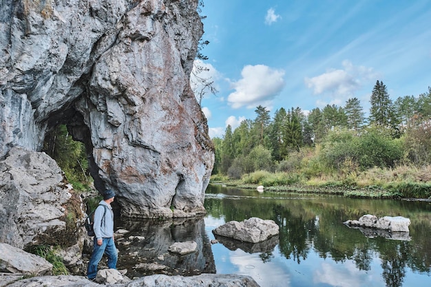 Senior male hiker standing on stony bank of river near rock Summer nature landscape