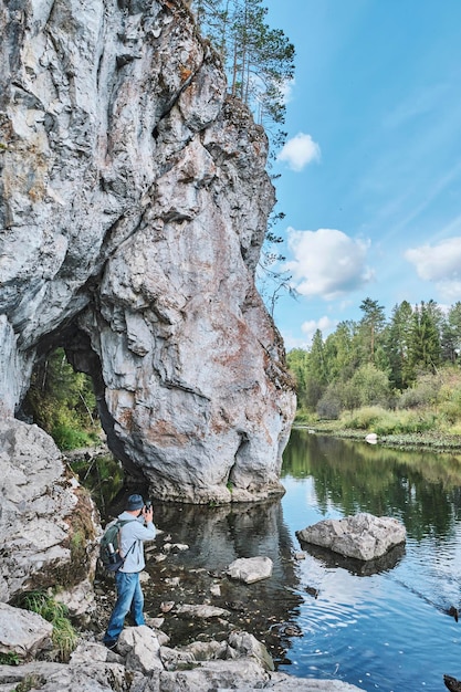 Senior male hiker taking pictures on mobile phone on bank of river near rock Summer nature landscape