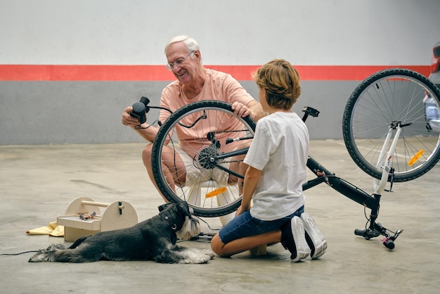 Senior male smiling and looking at pump while inflating tyre of bicycle helping grandson with repair on light parking with dog lying near