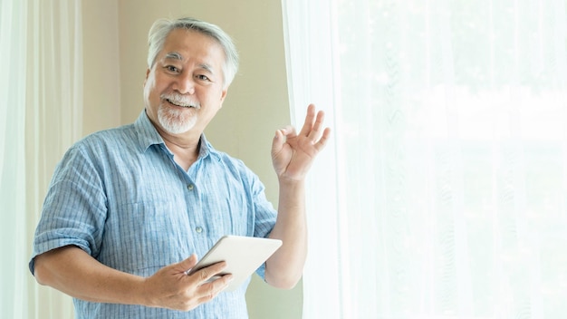 Senior Male using a smartphone and shows ok sign with hands tablet computer smiling feel happy in bedroom at home lifestyle senior concept