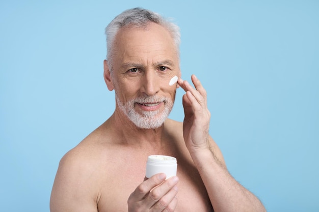 Senior man applying moisturizing nourishing anti aging cream from white a jar on blue background