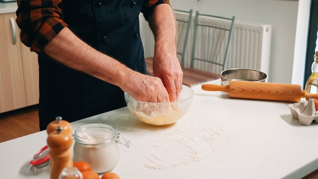 Senior man baker kneading bread dough in a glass bowl. Retired elderly chef with uniform sprinkling, sieving sifting raw ingredients by hand and mixing with flour for baking homemade pizza, cookies.