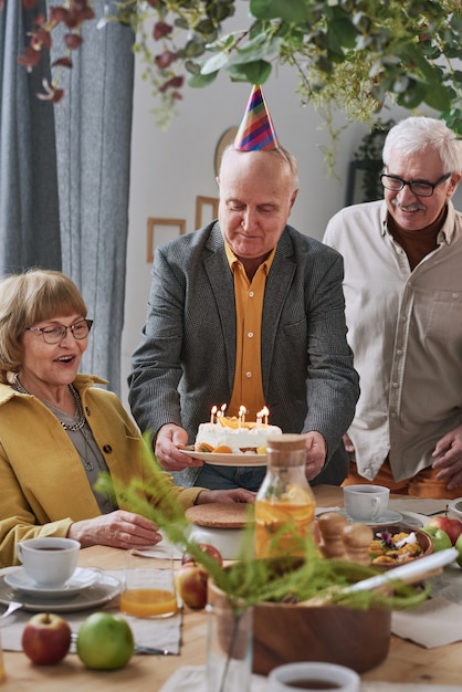 Senior man carrying cake with candles while they celebrating birthday of senior woman at the party