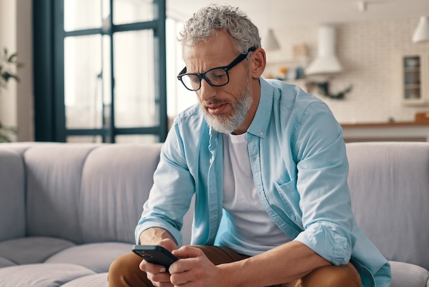 Senior man in casual clothing and eyeglasses using smart phone while sitting on the sofa at home