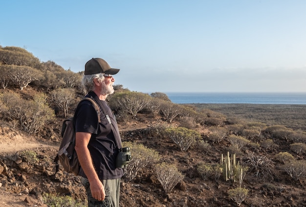 Senior man enjoying hiking in arid volcanic landscape. Looking at the horizon over sea. Backpack and binoculars, youthful elderly healthy lifestyle