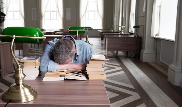 Senior man falls asleep over a textbook he is sitting at a table that is littered with teaching materials