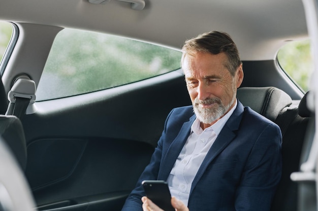 Senior man in formal wear sitting in a car looking at smartphone