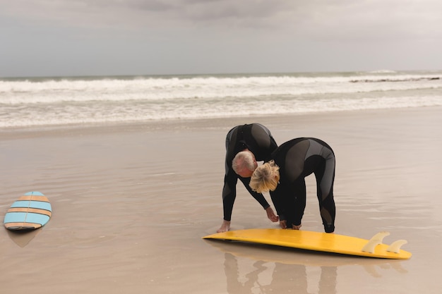 Photo senior man helping senior woman to wear surfboard leash on the beach