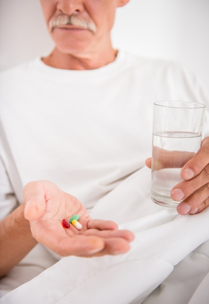 Senior man is drinking water with pills, sitting in bed.