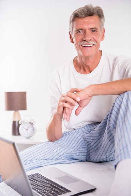 Senior man is using laptop while sitting in bed.