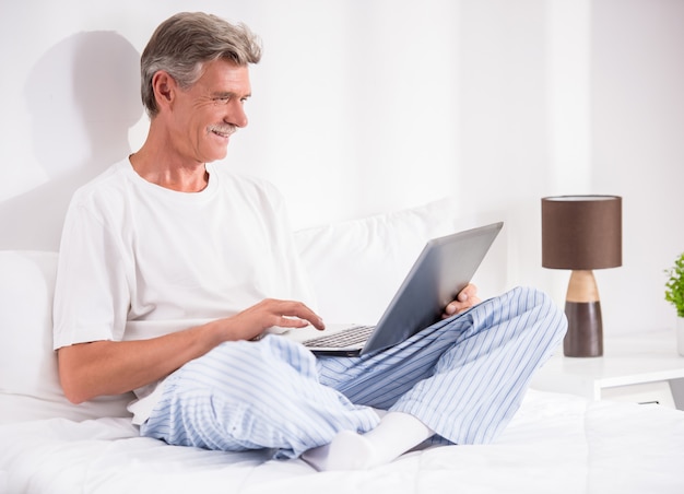 Senior man is using laptop while sitting in bed.