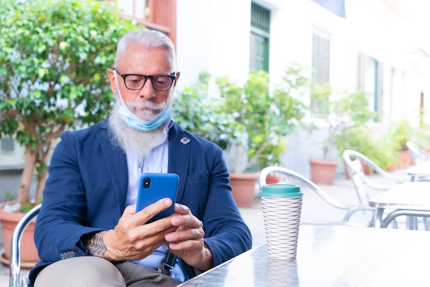 Senior man in mask with modern phone sitting in cafe during coronavirus outbreak, new normal concept