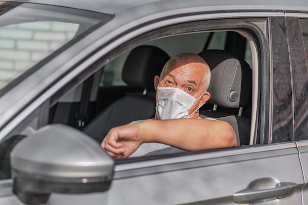 Senior man in medical mask driving a car, looking at the camera.