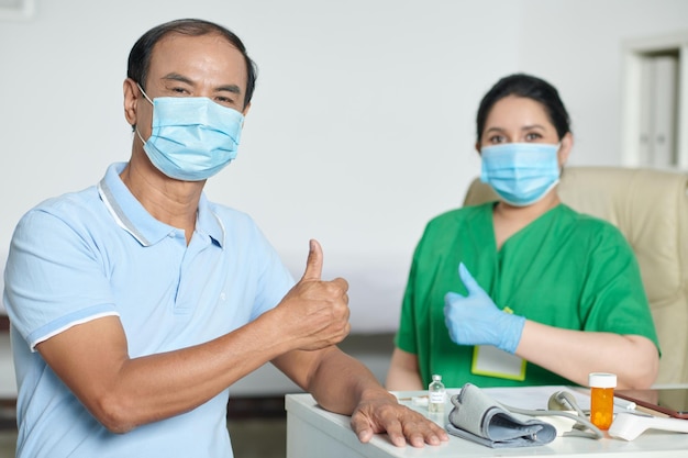 Senior man in medical mask showing thumbsup after successfully recovering from coronavirus