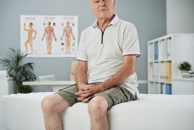 Senior man sitting on examination bed in medical office waiting for physiotherapist