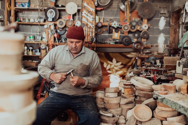 A senior man sitting in the workshop and processing wooden utensils in the old manual way.