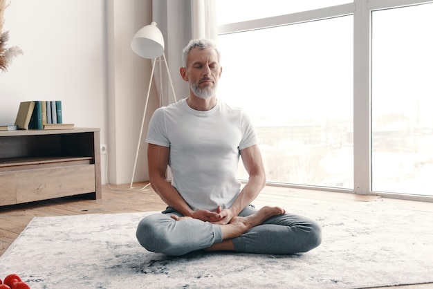 Senior man in sport clothing meditating while sitting on the floor in the lotus position at home