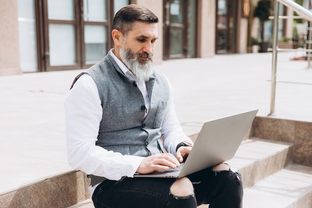 Senior man stylish man with gray beard spends time outdoors in laptop.