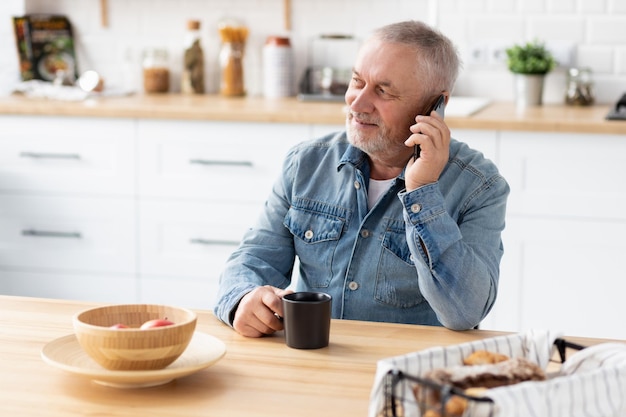 Senior man talking on phone sitting at the table in the home kitchen