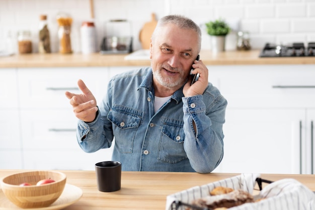 Senior man talking on phone sitting at the table in the home kitchen