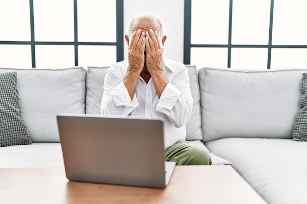 Senior man using laptop at home sitting on the sofa with sad expression covering face with hands while crying. depression concept.