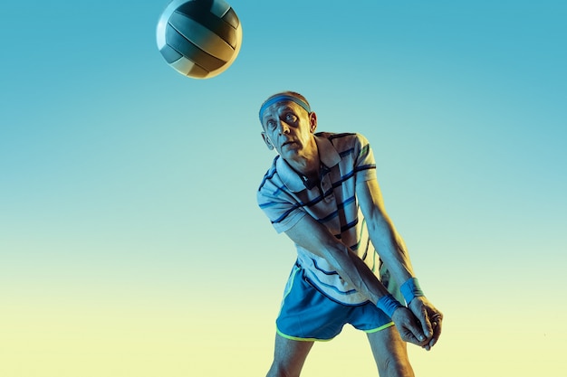 Senior man wearing sportwear playing volleyball on gradient background, neon light. Caucasian male model in great shape stays active. Concept of sport, activity, movement, wellbeing, confidence.