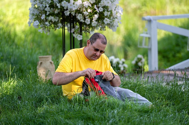 A senior man with a backpack sits on the grass in a park on a sunny day