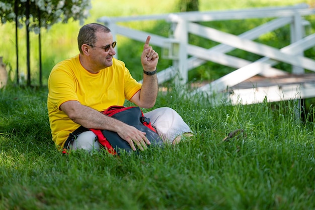 A senior man with a backpack sits on the grass in a park on a sunny day