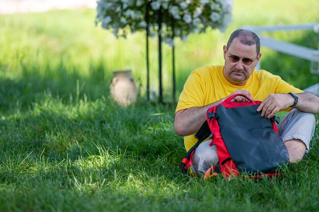 A senior man with a backpack sits on the grass in a park on a sunny day