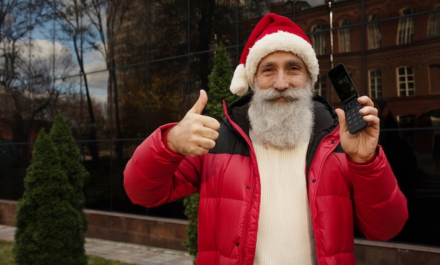 Senior man with beard in red santa hat presenting something outdoor. Xmas. New Year.