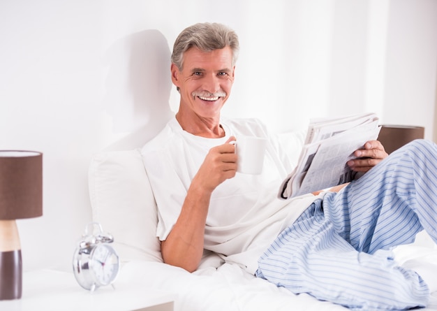 Senior man with a cup of coffee is reading newspaper in bed.