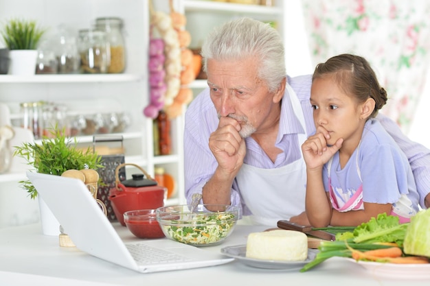 Senior man with granddaughter looking at laptop while preparing dinner in kitchen